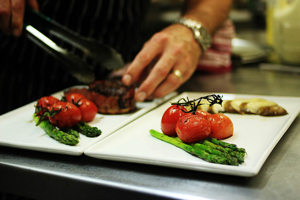 Man's hand plating steaks onto two square white plates with steamed whole tomatoes and asparagus on commercial, metal work surface. 