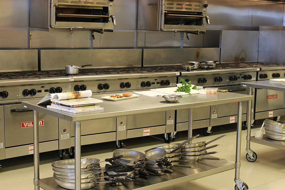 bank of stainless steel, wheeled commercial stoves and two overhead ovens behind two wheeled, stainless steel tables topped with ingredients and cookbooks. 