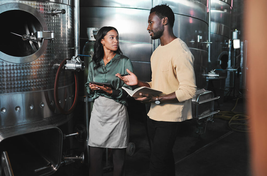 Young African American winemakers discussing production near equipment in winemaking facility. 