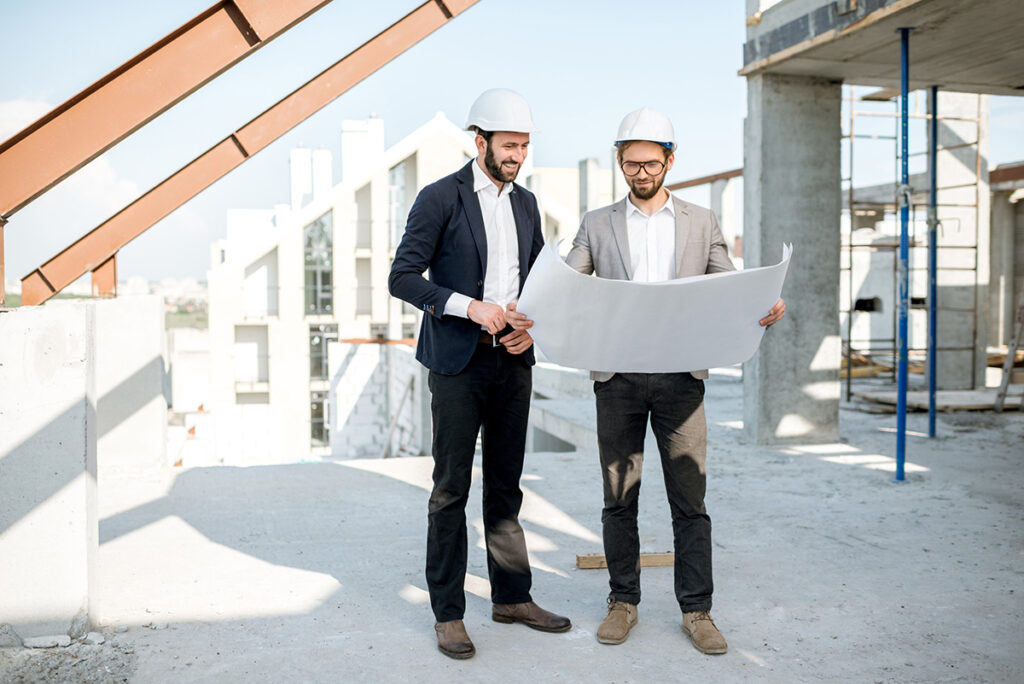Two commercial construction workers wearing suits and hard hats stand at a commercial construction site, looking over blueprints.