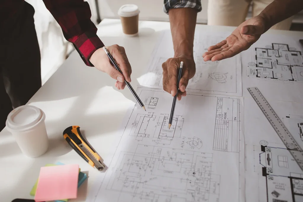 A commercial general contractor collaborates with engineers and architects to plan a commercial construction project, at a desk covered in printed plans and blueprints.