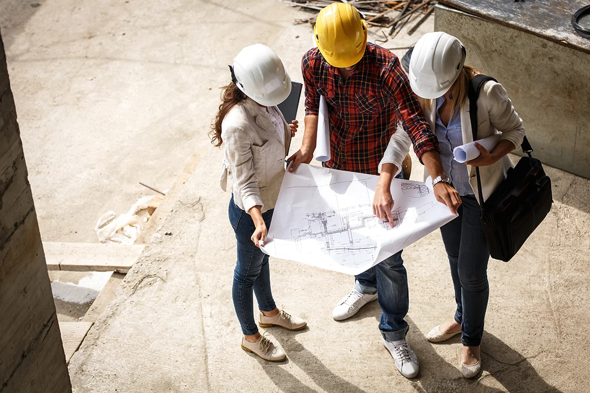 A commercial general contractor wearing a yellow hard hat and a red flannel stands at a commercial construction site, going over blueprints with two other construction professionals in white hard hats.