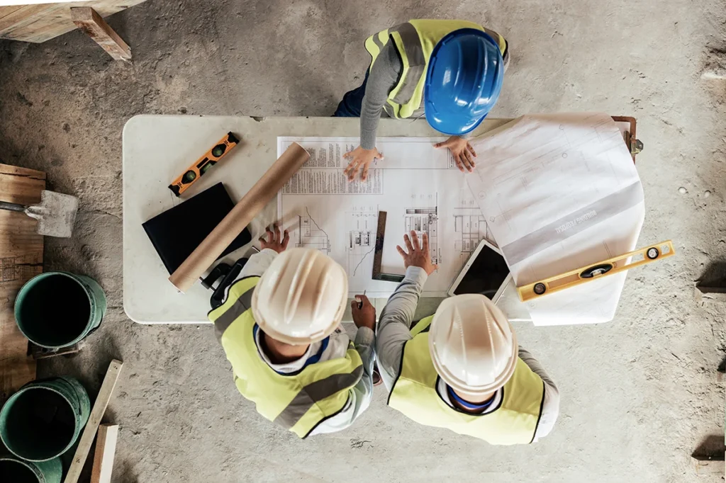 A design-build team wearing hard hats stands over a table with blueprints and construction plans, showing the concept of the benefits of including a general contractor in the design phase