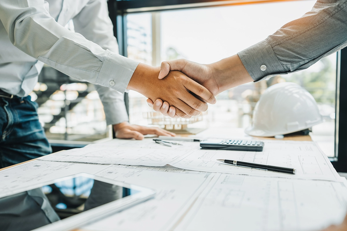 A tenant improvement contractor shaking hands with a client over a desk, covered in blueprints and construction plans